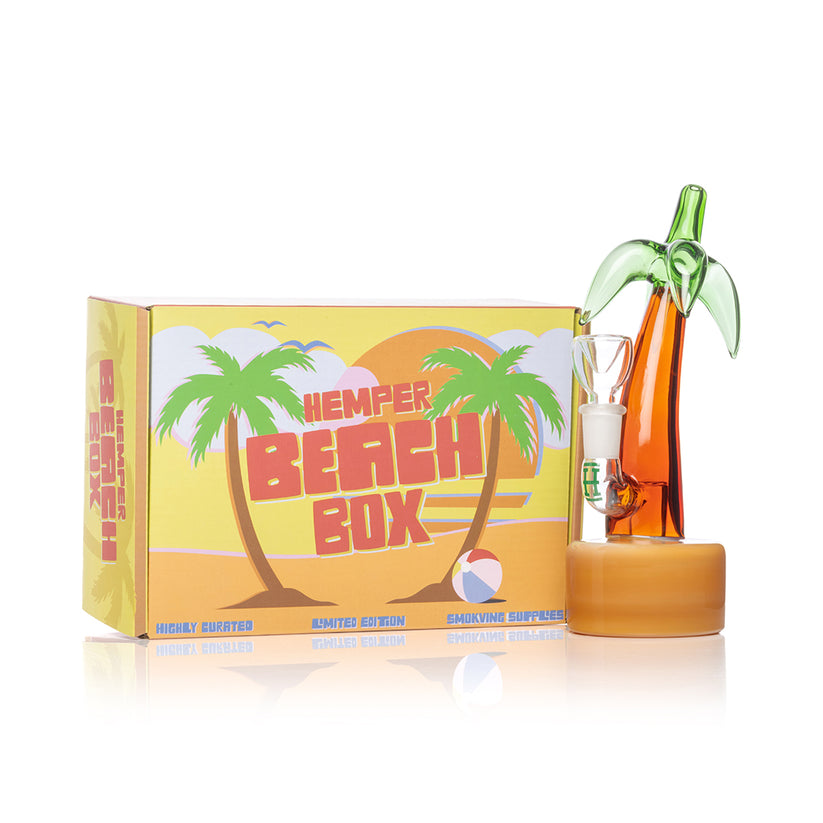 Front view of Hemper 7-inch Palm Tree water pipe next to yellow Beach Box, ready-for-gifting palm-themed glass setup.