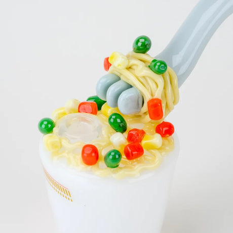 Overhead detail of sculpted noodles and veggies atop the cup, showing the curved fork mouthpiece entering the lid.