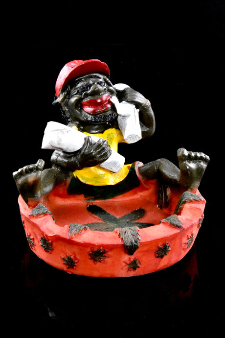 Close-up of red Rasta resin ashtray with figure reclining across rim, marijuana leaf motifs around base, dramatic black background.