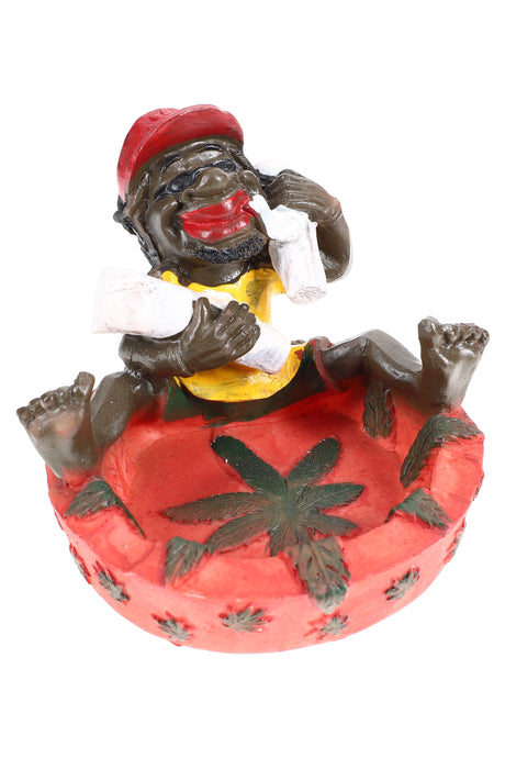 Playful Rasta resin ashtray with red bowl and relaxed figure hugging giant joints, four-inch diameter, studio white backdrop.