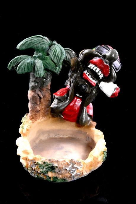 Beach-themed resin ashtray with leaning Rasta under palm, worn stone-look bowl and green foliage accents, dark studio lighting.