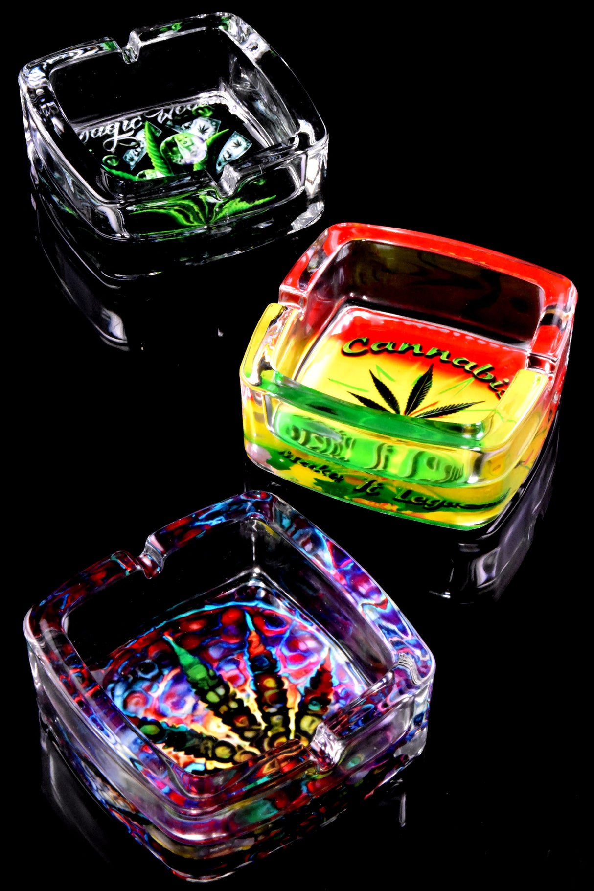 Square glass ashtray trio bearing cannabis leaf graphics in green Rasta gradient and tie-dye swirls, black stage.