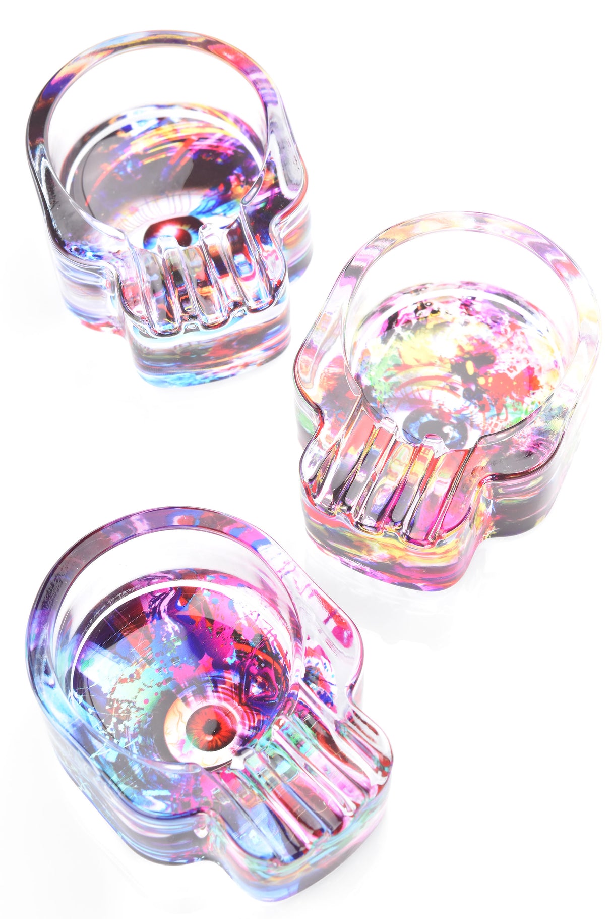 Skull glass ashtray set with psychedelic splatter and eyeball graphics, iridescent hues, deep circular bowl and front grooves on white backdrop.