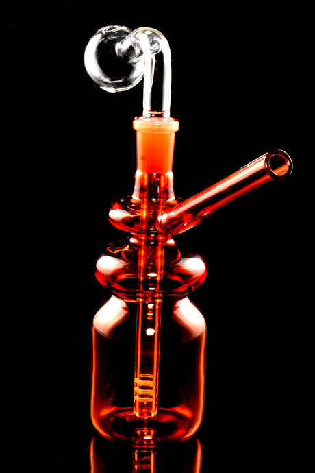 Orange bottle oil burner rig highlighting clear burner dome and snug glass-on-glass connection, studio black background.