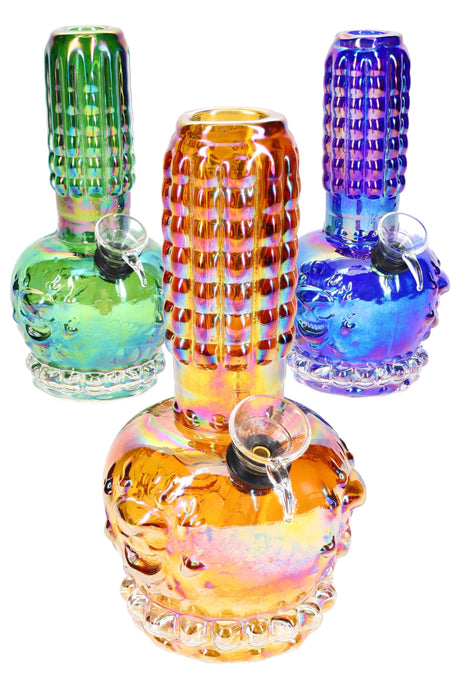 Rainbow metallic 7-inch beaker bong with sculpted skull base, ribbed grip neck, and grommet bowl, shown in three assorted colors on white background.
