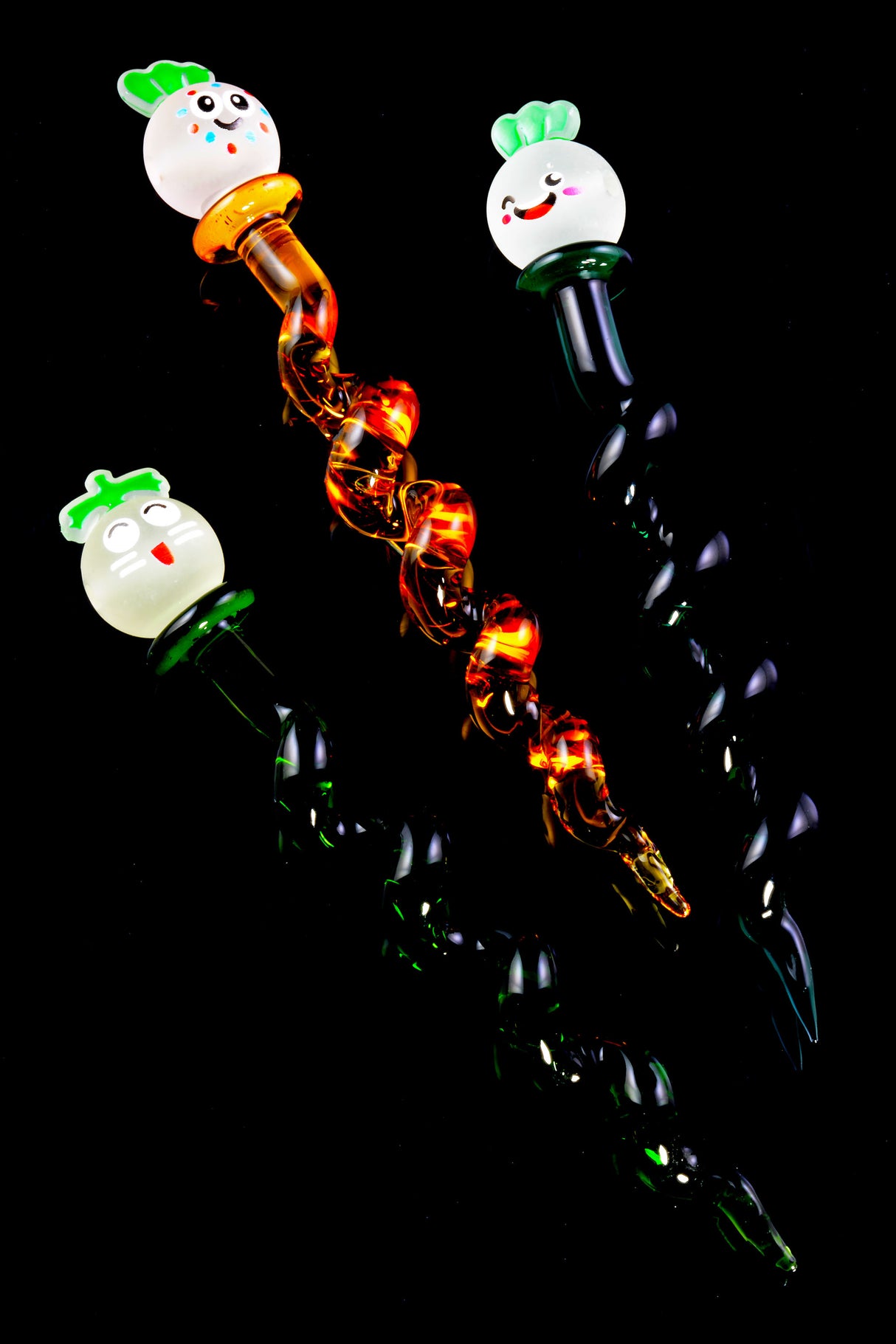 Trio of twisted glass dabbers featuring happy radish faces and emerald, amber, and black spiral stems, arranged diagonally on deep black background for contrast.