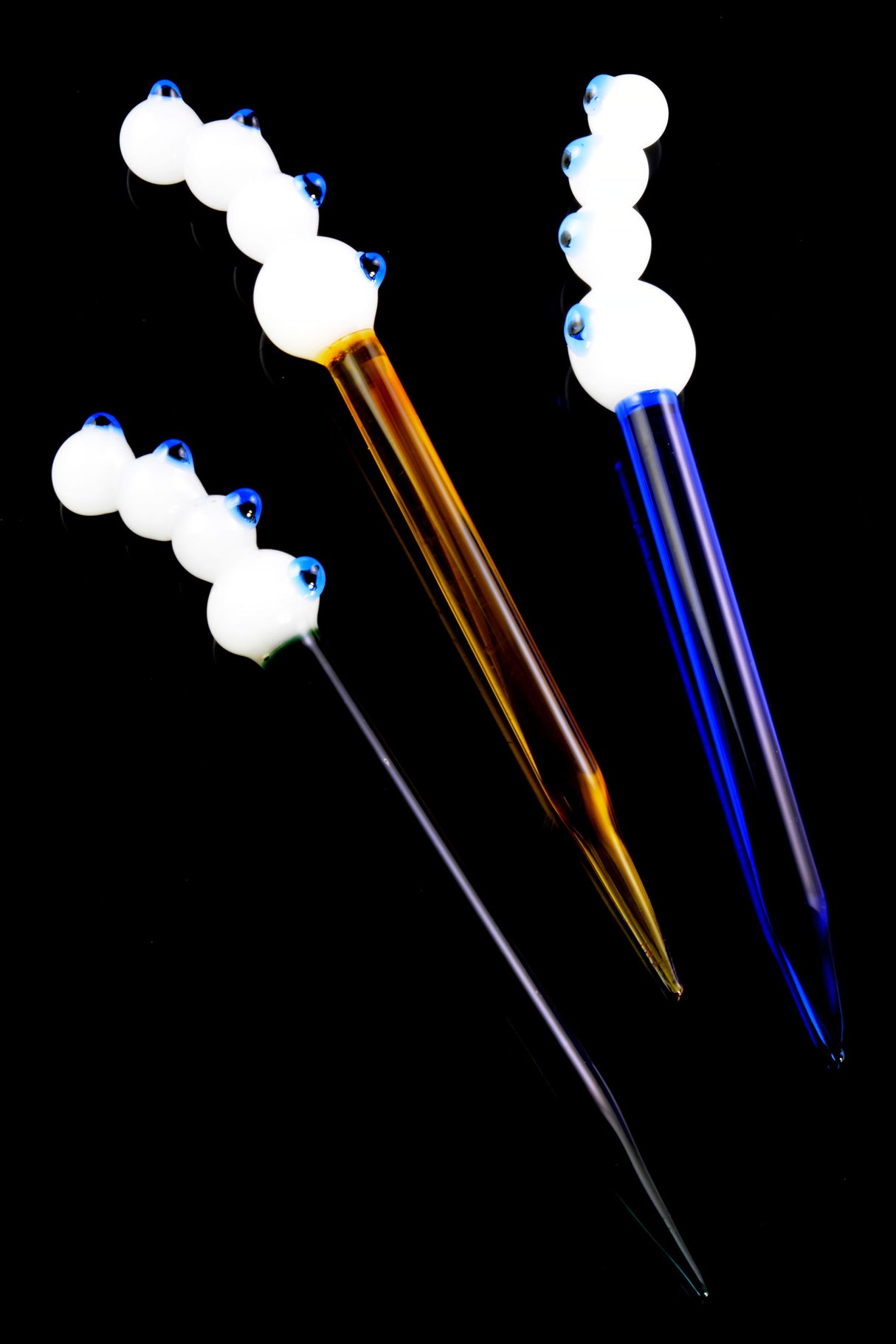 Three four-eyes glass dab tools—amber, purple, and clear stems—with raised white marble grips and cobalt pupils, arranged on black backdrop for vivid contrast.