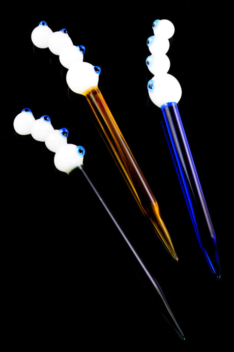 Three four-eyes glass dab tools—amber, purple, and clear stems—with raised white marble grips and cobalt pupils, arranged on black backdrop for vivid contrast.