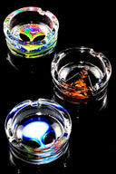 Trio of tempered glass ashtrays featuring alien and abstract artwork, side-by-side on reflective black surface.