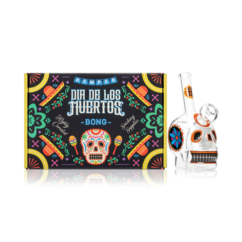 Front view of Dia De Los Muertos skull mini glass bong next to matching Hemper gift box, showing bright sugar-skull graphics.