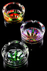 3.25-inch tempered glass ashtray with multicolor cannabis leaf graphic, angled overhead on glossy black background.