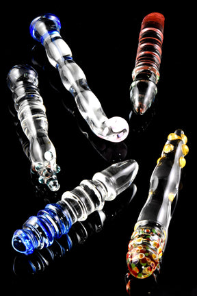 Assorted US-made large glass dildos with ridges, bulbs, and swirled accents, clear with colored details, arranged on a glossy black surface.