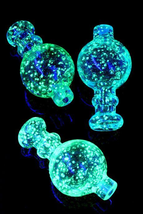 Group of frit bubble carb caps glowing bright under UV/blacklight, showing speckled effect and rounded handles; assorted colors.