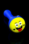 Blue silicone pipe with yellow face cap showing tongue out; clear removable glass bowl in the top.