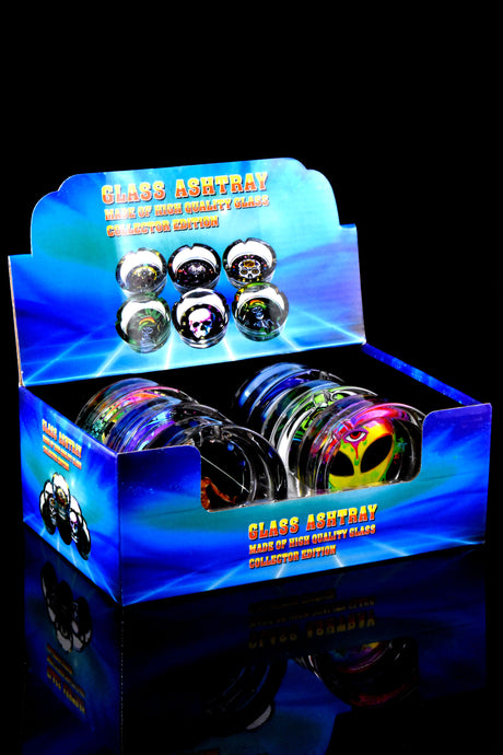 Retail display box filled with assorted pop-design tempered glass ashtrays, front-left angle showing stacked units.