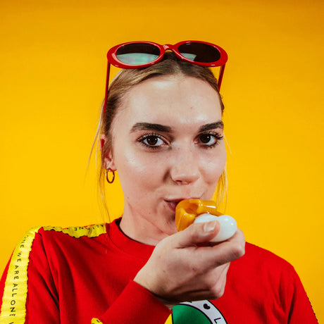 Person holding Goody Glass Bananza hand pipe with yellow backdrop and red sunglasses visible