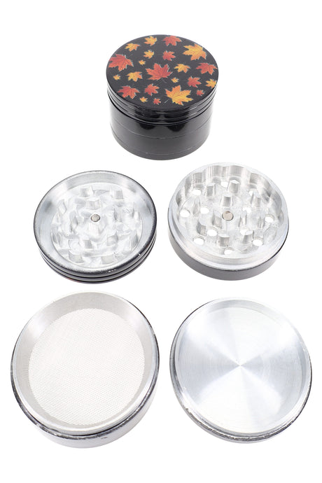 Black 56 mm 4-part aluminum herb grinder with fall-leaf decal lid, shown disassembled with teeth, mesh sifter and catch.