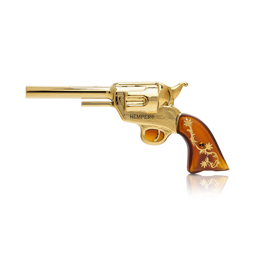 Side profile of Hemper Peacemaker gold revolver glass hand pipe with amber scrollwork grip and long barrel on white.