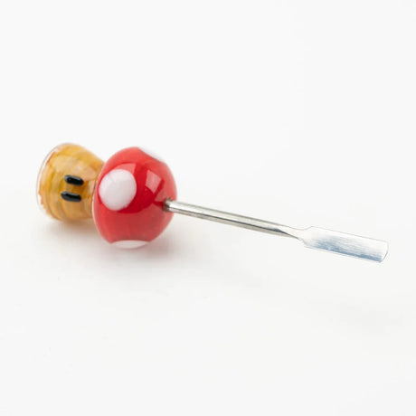 Angled shot of mushroom glass dab tool lying down, focusing on weighted base, red cap, and metal spatula tip against white studio surface.