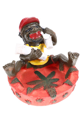Playful Rasta resin ashtray with red bowl and relaxed figure hugging giant joints, four-inch diameter, studio white backdrop.