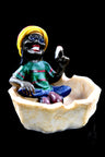Wide resin ashtray shaped like seashell with chill Rasta wearing striped hat, holding joint above cream-textured basin, black background.
