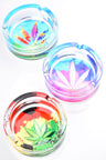 Round 4-inch glass ashtrays sporting pastel cannabis leaf graphics in aqua, purple and rasta gradients; deep bowl, three grooves, studio overhead lighting.