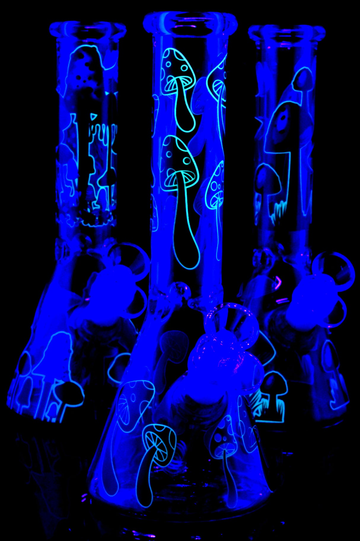 Trio of glow mushroom beaker bongs under UV light highlighting neon outlines; ice pinch and glass-on-glass joints visible, assorted graphics.