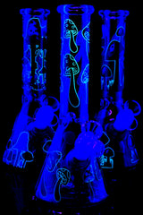 Trio of glow mushroom beaker bongs under UV light highlighting neon outlines; ice pinch and glass-on-glass joints visible, assorted graphics.