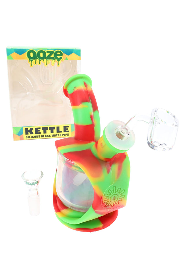 Rasta swirl silicone glass water pipe with quartz banger attached and boxed packaging behind, compact 6″ bubbler design.