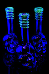 Three metallic skull soft-glass bongs glowing bright green frit rings under UV light, showcasing sculpted bases and reflective finish against dark background.