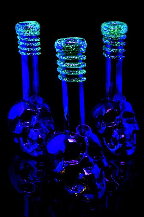 Three metallic skull soft-glass bongs glowing bright green frit rings under UV light, showcasing sculpted bases and reflective finish against dark background.