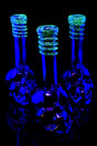 Three metallic skull soft-glass bongs glowing bright green frit rings under UV light, showcasing sculpted bases and reflective finish against dark background.