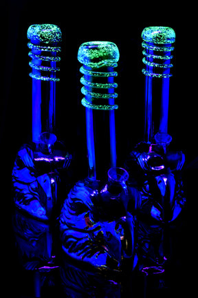 Metallic eagle water pipes glowing bright green frit rings under UV light, showcasing reflective bases in dark room.