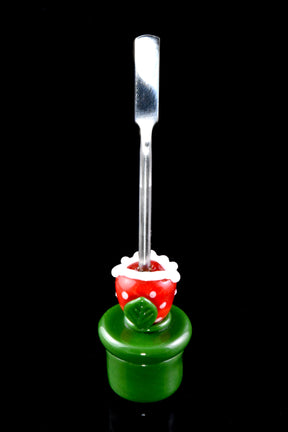 Piranha-plant glass dab tool with stainless flat scoop tip, red polka-dot head and green pipe base, standing upright on black background.