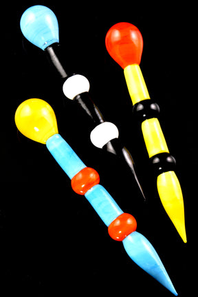 Thick 5.5-inch glass dab tools with bold red-yellow-black and sky-blue color blocks, bulbous handle tops, and tapered points, arranged on black backdrop.