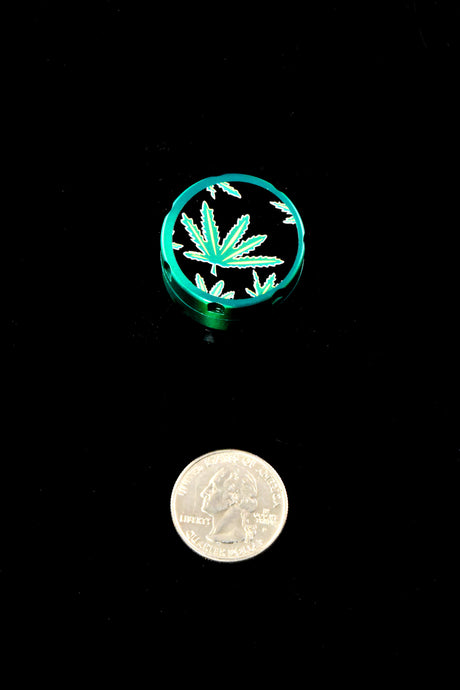 Teal mini herb grinder with leaf-decal lid shown beside a quarter for scale; ultra-compact pocket grinder.