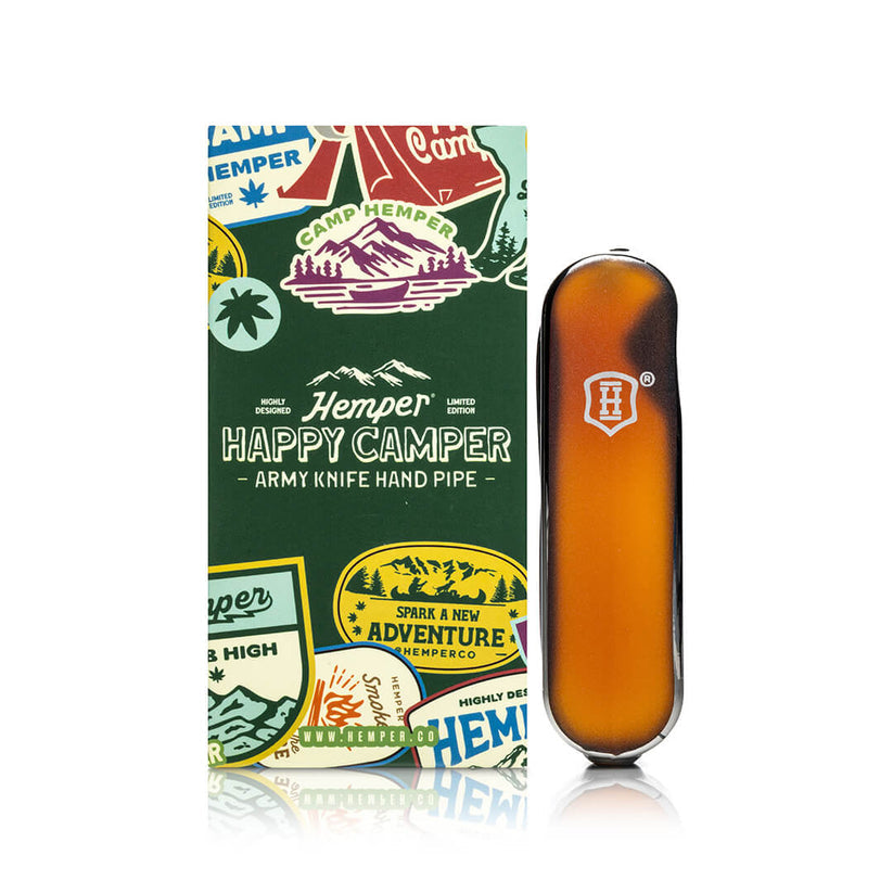 Front view of amber Happy Camper army knife glass hand pipe standing beside sticker-covered Camp Hemper box for hero shot.